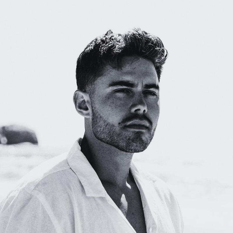 Black-and-white headshot of a young man with a beard, wearing a white shirt, with a serious expression and ocean backdrop.